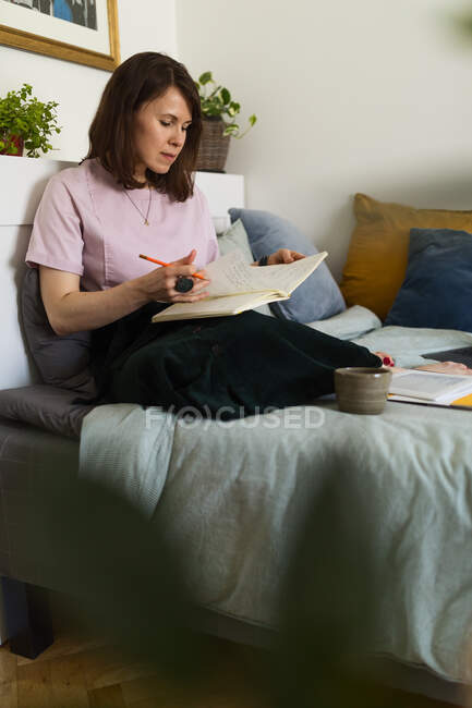 Designer gráfica feminina sentada na cama com caderno de esboços e trabalhando em projeto remoto em casa — Fotografia de Stock