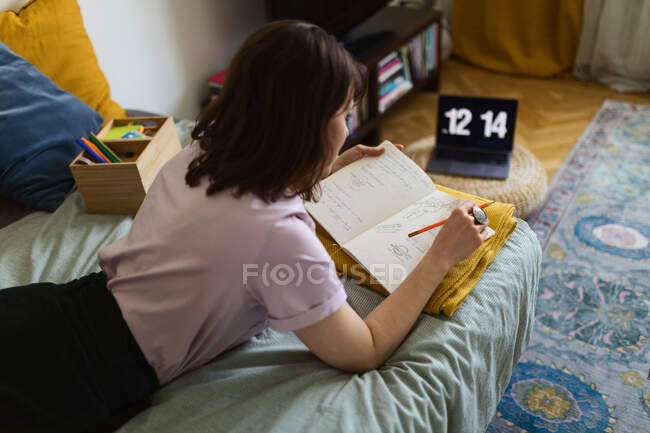 Vista lateral del diseñador gráfico femenino acostado en la cama con cuaderno de bocetos y trabajando en un proyecto remoto en casa - foto de stock