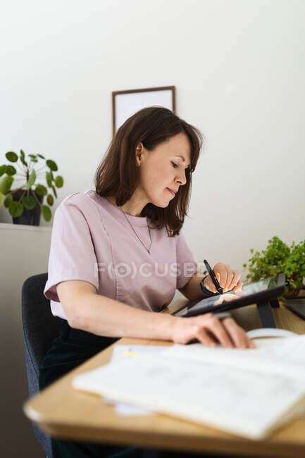 Vista lateral de la mujer dibujo de la imagen en la tableta gráfica mientras está sentado en la mesa en la oficina en casa - foto de stock