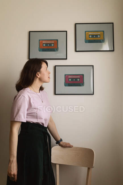 Adulto feminino inclinado na cadeira e admirando imagens emolduradas de cassetes penduradas na parede em casa — Fotografia de Stock
