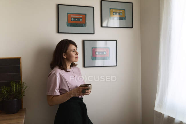 Dreamy adult woman with cup of hot drink looking out window and relaxing while leaning on wall near framed images of cassettes at home — Stock Photo