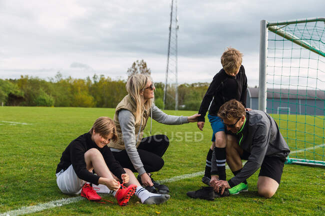Родители помогают сыновьям-подросткам надевать футбольные сапоги во время подготовки к игре в футбол на зеленом поле — стоковое фото