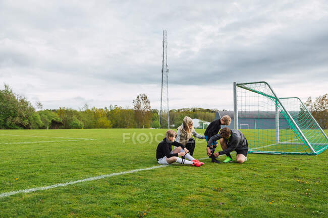 Pais ajudando filhos adolescentes a colocar botas de futebol enquanto se preparam para jogar futebol no campo verde — Fotografia de Stock