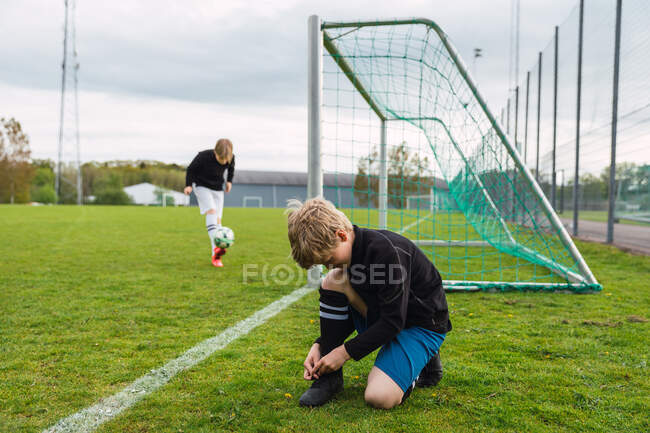 Vista laterale del ragazzo adolescente che lega i lacci sulle scarpe da calcio mentre si prepara per l'allenamento sul campo — Foto stock