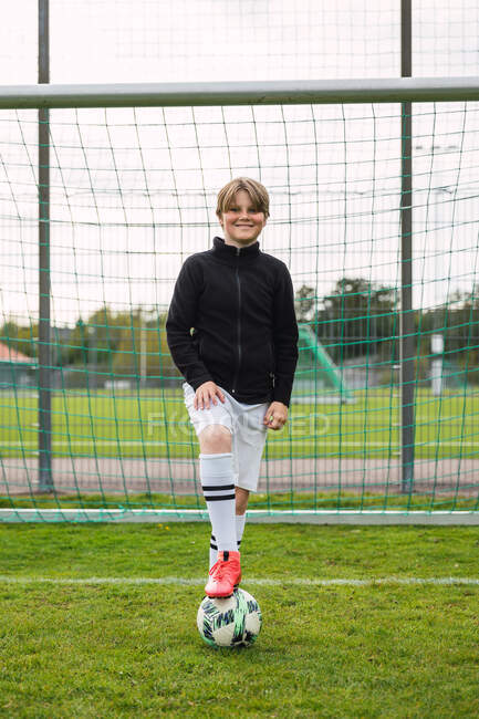 Deleitado adolescente en ropa deportiva de pie con pelota en el campo de fútbol cerca de la red y mirando a la cámara - foto de stock