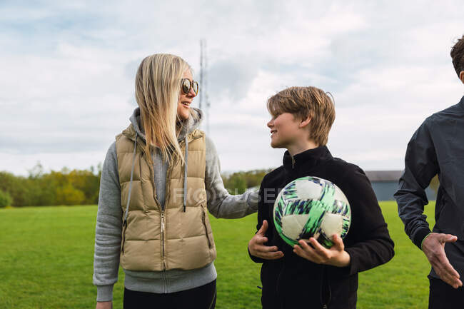 Веселая мать и сын-подросток в спортивной одежде собираются на зеленом футбольном поле за совместную игру в футбол на выходных — стоковое фото