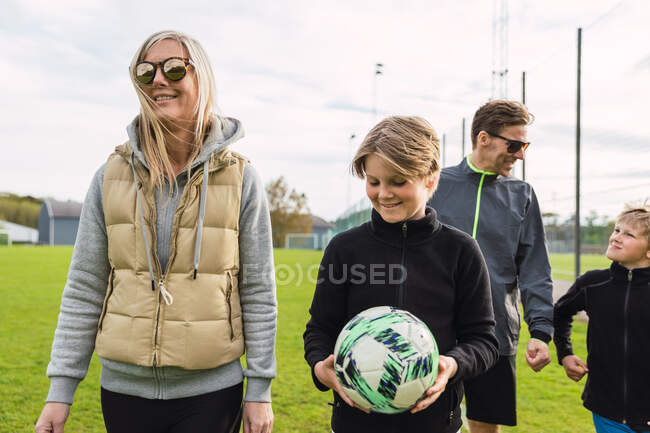 Веселая пара и сыновья-подростки в спортивной одежде собираются на зеленом футбольном поле, чтобы вместе играть в футбол на выходных — стоковое фото