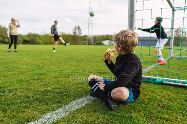 Adolescente irreconocible sentado en el campo de fútbol y comiendo plátano mientras mira a la familia jugando fútbol - foto de stock