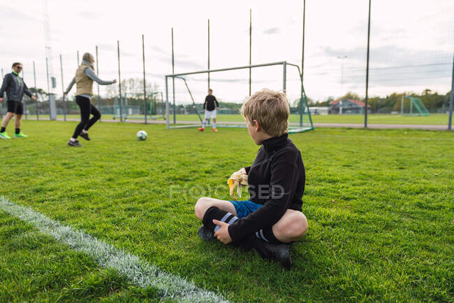 Невпізнаваний хлопчик сидить у футбольному полі і їсть банан, дивлячись на те, як сім'я грає у футбол — стокове фото