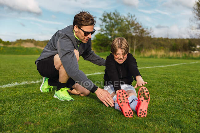 Adolescente niño en ropa deportiva piernas estiradas antes del entrenamiento de fútbol con la ayuda de entrenador masculino en el campo verde - foto de stock