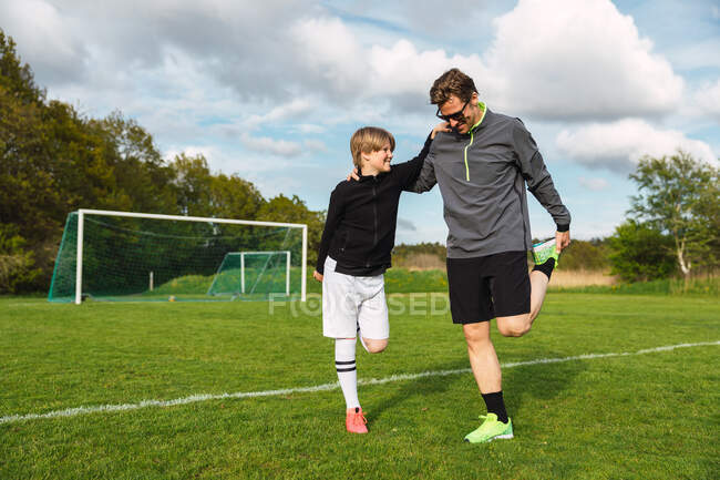 Alegre padre y adolescente en ropa deportiva que estira las piernas mientras se prepara para jugar fútbol en el campo de fútbol en verano - foto de stock