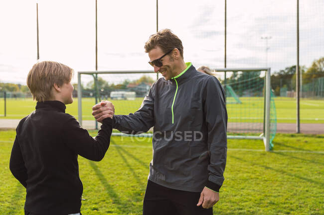 Vista lateral del padre sonriente y el hijo adolescente en ropa deportiva estrechando la mano y saludándose durante el entrenamiento de fútbol en el campo - foto de stock