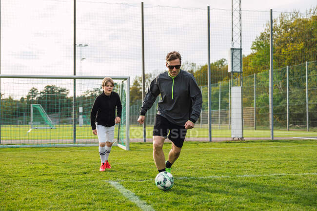 Fröhlicher Vater und Teenager-Sohn in Aktivkleidung spielen Fußball, während sie Ball kicken und über das Feld rennen — Stockfoto
