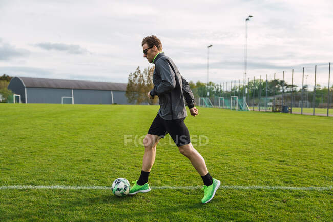 Вид сбоку на футболиста в спортивной форме, пинающего мяч во время тренировки на зеленом футбольном поле — стоковое фото