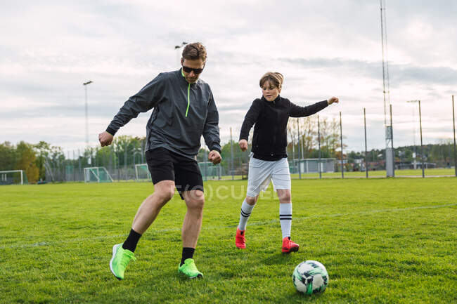 Cheerful father and teenage son in activewear playing football while kicking ball and running along field — Stock Photo