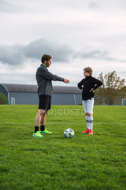 Alegre padre e hijo adolescente en ropa deportiva jugando al fútbol mientras patea la pelota y corre por el campo - foto de stock
