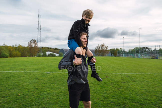 Веселый отец носит мальчика-подростка на плече, стоя на футбольном поле — стоковое фото