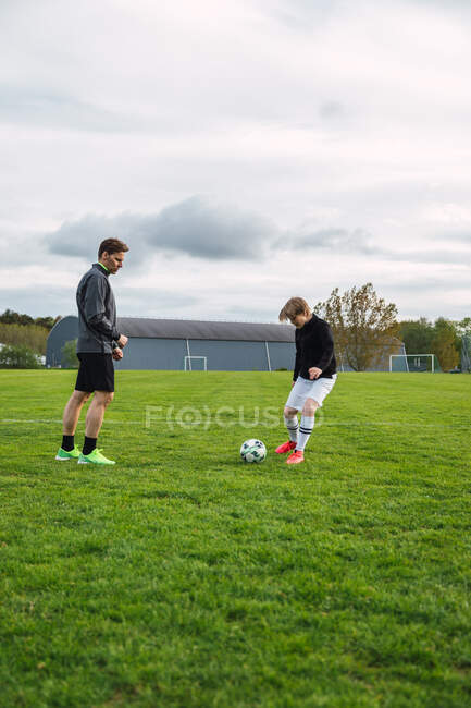 Alegre padre e hijo adolescente en ropa deportiva jugando al fútbol mientras patea la pelota y corre por el campo - foto de stock