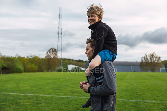Cheerful father carrying teenage boy on shoulder while standing on football field — Stock Photo