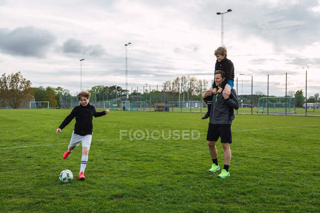 Cheerful teenage boy kicking ball and playing football in field with father and brother during weekend — Stock Photo
