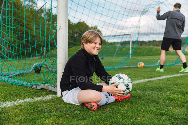Вид сбоку положительного футболиста-подростка в спортивной форме, сидящего с мячом на зеленом поле во время тренировки — стоковое фото