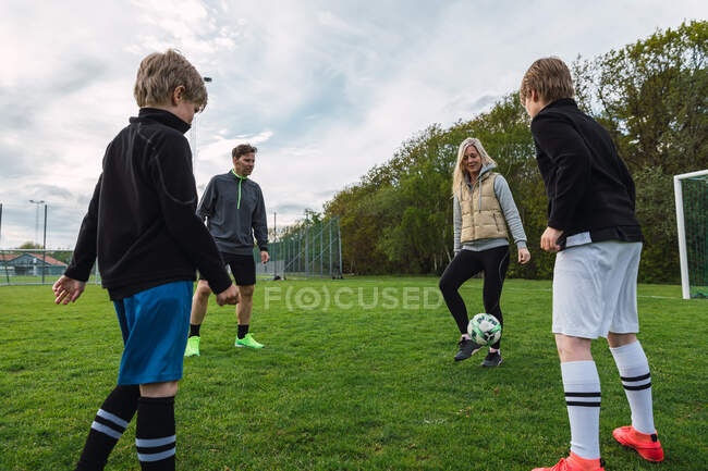 Счастливые спортивная пара и подростки играют в футбол вместе, пиная мяч в зеленом поле летом — стоковое фото