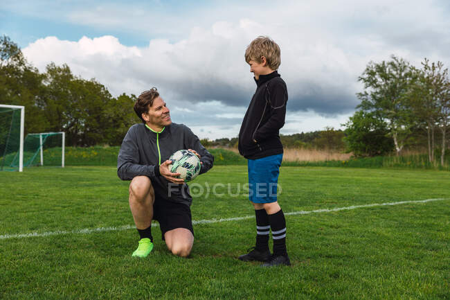 Улыбающийся отец с мячом смотрит на сына-подростка в спортивной одежде, проводя время вместе на футбольном поле в выходные — стоковое фото