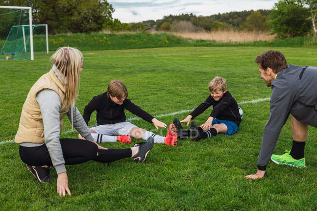 Веселая семья в спортивной одежде растягивает ноги на зеленом поле и готовится к совместной игре в футбол — стоковое фото