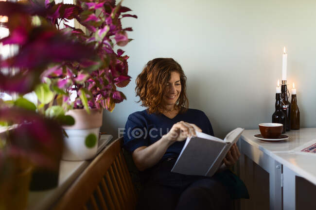 Adult female reading interesting book while sitting at table decorated with burning candles at home — Stock Photo