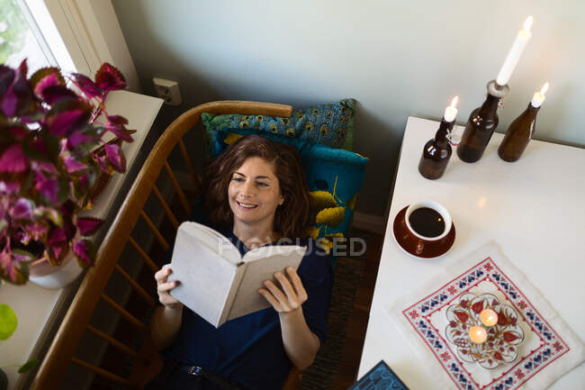 Adulte femelle lecture intéressant livre tout allongé à la table décoré avec des bougies brûlantes à la maison — Photo de stock
