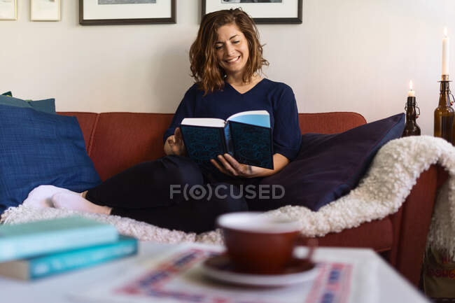 Felice femmina guardando lontano e sognando mentre seduto sul divano e leggendo libro a casa — Foto stock