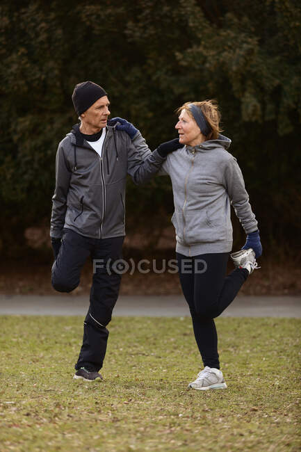 Full body of old couple wearing sportswear warming up before training and helping each other in park — Stock Photo