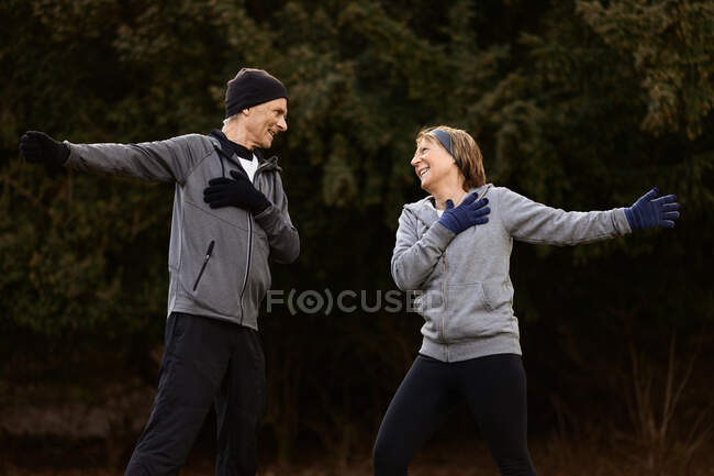 Позитивна стара пара носить спортивний одяг, розтягуючи руки під час тренувань у парку і дивлячись один на одного — стокове фото