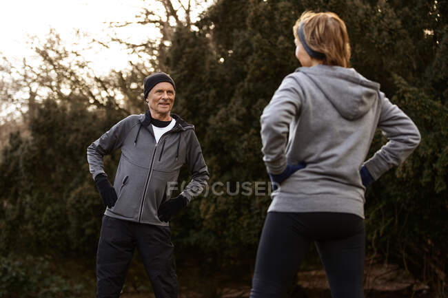 Позитивная пожилая пара в спортивной одежде, вытягивающая руки во время тренировок в парке и смотрящая друг на друга — стоковое фото