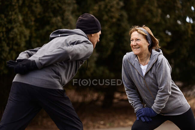 Positivo casal velho vestindo roupas esportivas esticando as pernas enquanto se exercitam no parque e olhando um para o outro — Fotografia de Stock