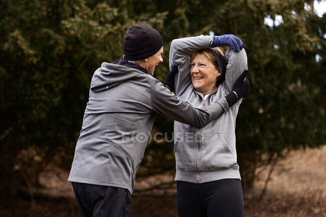 Mujer mayor sonriente levantando las manos mientras hace ejercicio de calentamiento con el apoyo del hombre durante el entrenamiento en el parque - foto de stock