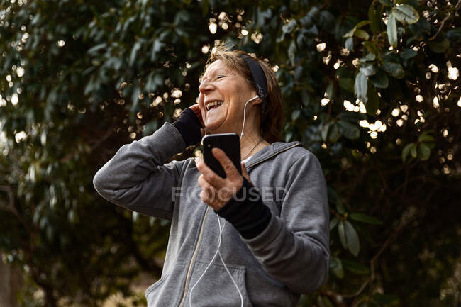 Angle bas de la vieille femme en vêtements de sport écouter de la musique dans les écouteurs pendant l'entraînement en plein air — Photo de stock