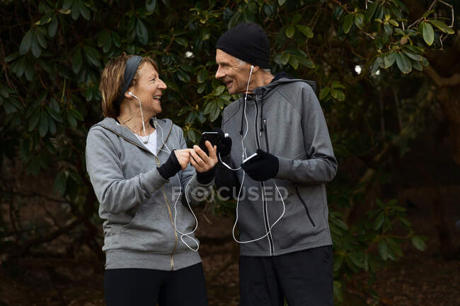 Улыбающаяся пожилая пара в спортивной одежде и наушниках, стоящая вместе под ветками деревьев и обменивающаяся мобильным телефоном во время тренировки — стоковое фото