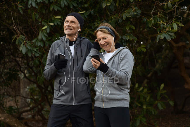 Smiling senior couple in sportswear and earphones standing together under tree branches and sharing mobile phone during fitness workout — Stock Photo