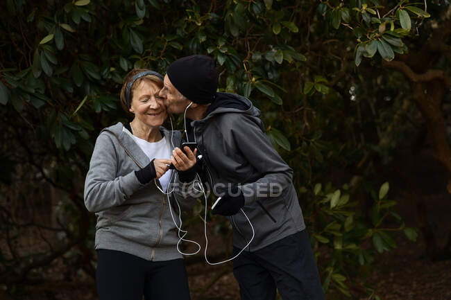 Lächelndes Senioren-Paar in Sportkleidung und Kopfhörern, das zusammen unter Ästen steht und beim Fitnesstraining sein Handy austauscht — Stockfoto