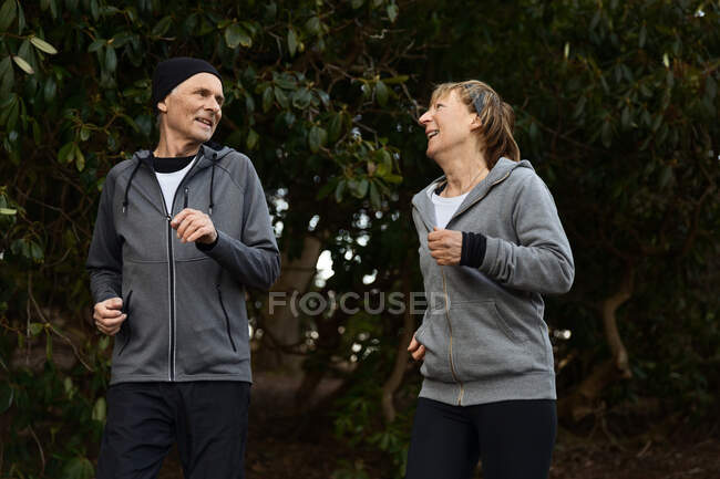 Улыбающаяся пожилая пара в спортивной одежде и пробежка между зелеными кустами в парке во время фитнес-тренировок — стоковое фото