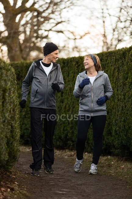 Corpo inteiro de casal idoso sorridente vestindo roupas esportivas e luvas e correndo entre arbustos verdes no parque durante o treinamento de fitness — Fotografia de Stock