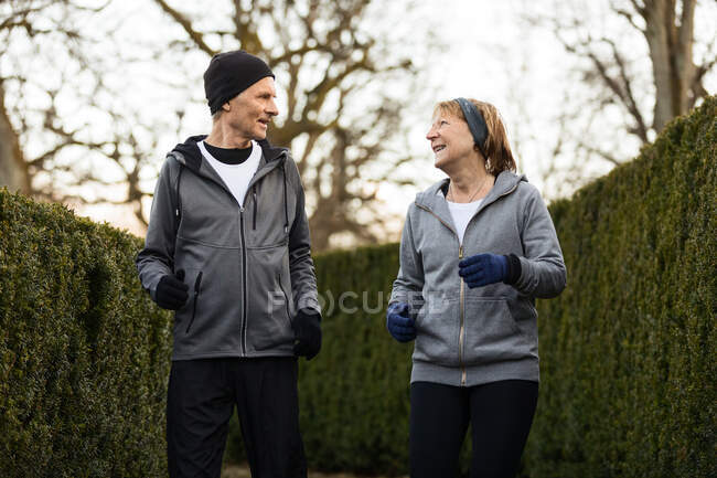 Pareja anciana sonriente que usa ropa deportiva y guantes y trota entre arbustos verdes en el parque durante el entrenamiento de fitness - foto de stock