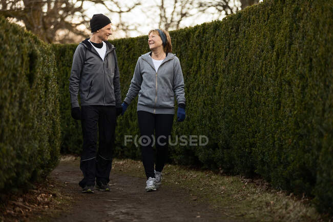 Corps entier d'un couple d'âge souriant portant des vêtements de sport et des gants et marchant entre des buissons verts dans le parc pendant l'entraînement de fitness — Photo de stock