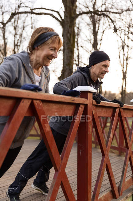 Sorridente donna anziana che fa spinta dal corrimano in legno con il partner mentre fa allenamento all'aperto nel parco autunnale — Foto stock