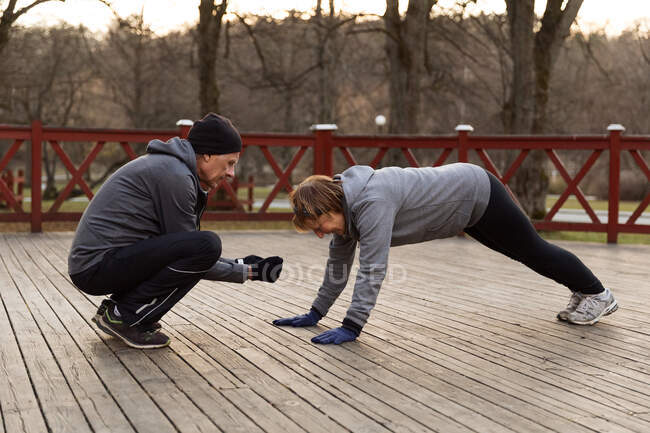 Full body side view of man assisting old woman doing high plank exercise in park during fitness training — Stock Photo
