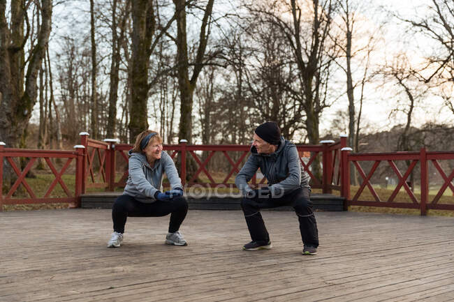 Full body of smiling senior couple doing squats during fitness session in park among leafless trees and looking at each other — Stock Photo