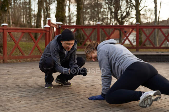 Повне тіло чоловіка, який допомагає старій жінці робити вправи на високій дошці в парку під час тренувань з фітнесу — стокове фото