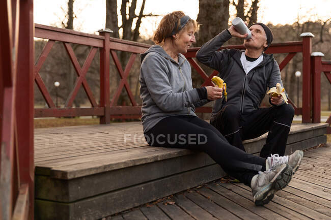 Cuerpo completo de pareja mayor bebiendo agua y comiendo plátanos mientras se sienta en el podio de madera después del entrenamiento - foto de stock
