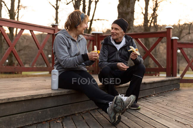Corpo inteiro de casal de idosos tendo pausa no treinamento e comer alimentos saudáveis enquanto conversam uns com os outros — Fotografia de Stock
